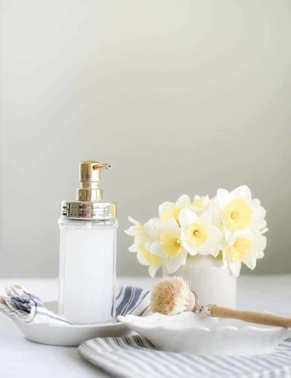 Liquid hand soap in glass jar pictured with daffodils and wooden scrubbing brush
