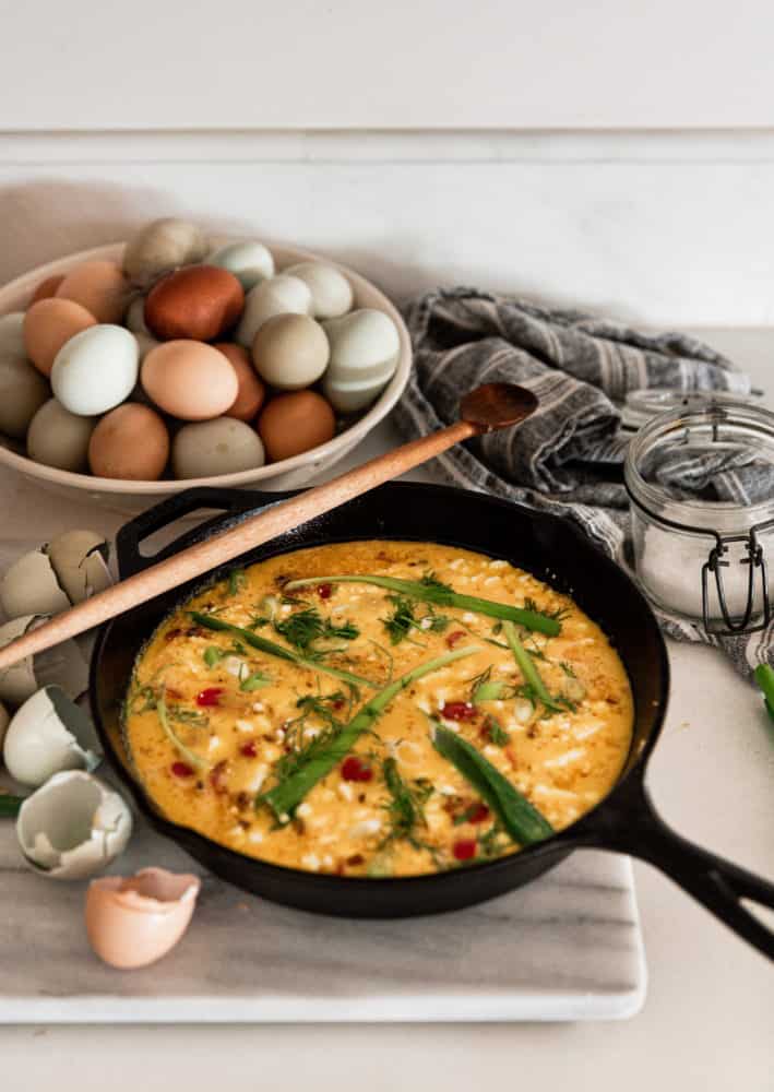 Uncooked frittata on a marble cutting board in a cast iron skillet, with dish of farm fresh eggs in the upper left corner.