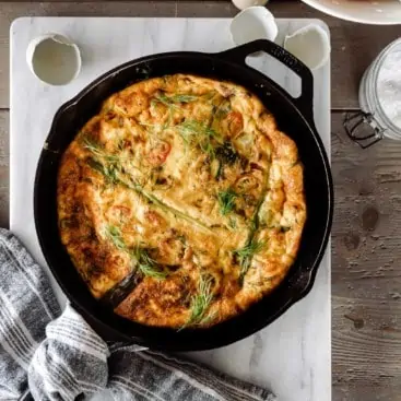 Baked frittata in a cast iron skillet on a marble board with farm fresh eggs next to it.