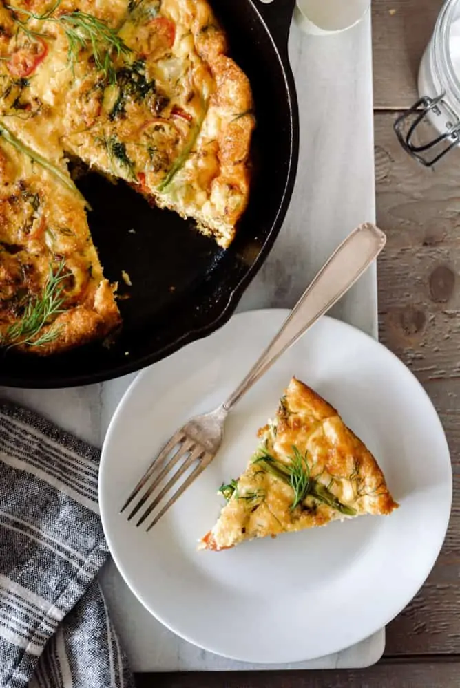 Baked frittata in a cast iron skillet on a marble board with slice on a plate with a fork.