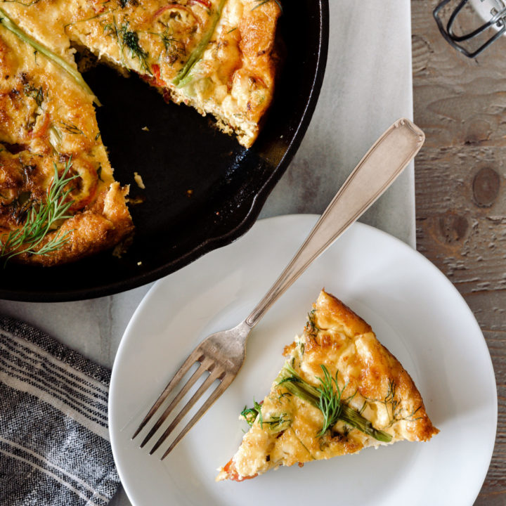 Baked frittata in a cast iron skillet on a marble board with slice on a plate with a fork.