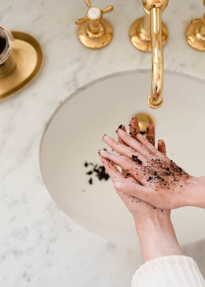 Woman using DIY coconut coffee scrub on hands over marble sink with brass faucet.