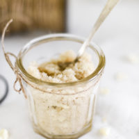 Glass jar with homemade sugar scrub and silver spoon on table with white linen tablecloth.