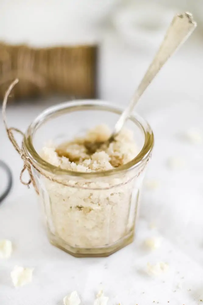 Glass jar with homemade sugar scrub and silver spoon on table with white linen tablecloth.