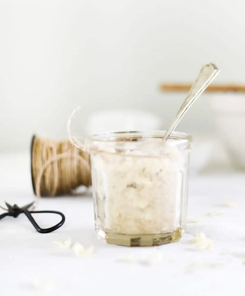 Glass jar with homemade sugar scrub and silver spoon on table with white linen tablecloth.