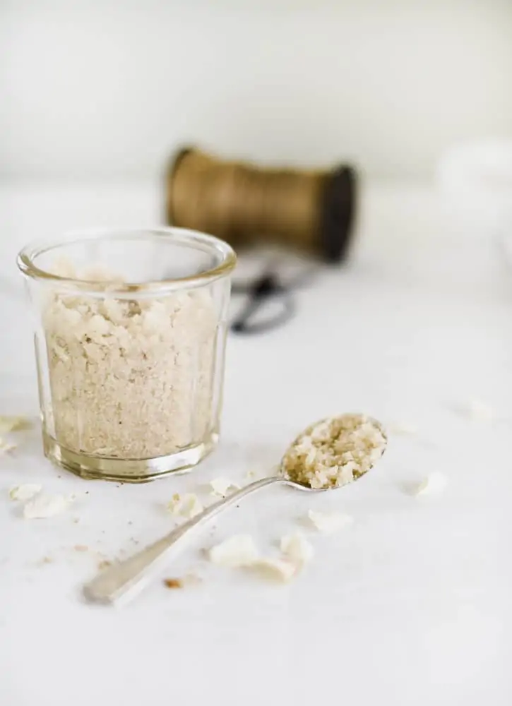 Glass jar with homemade sugar scrub and silver spoon on table with white linen tablecloth.