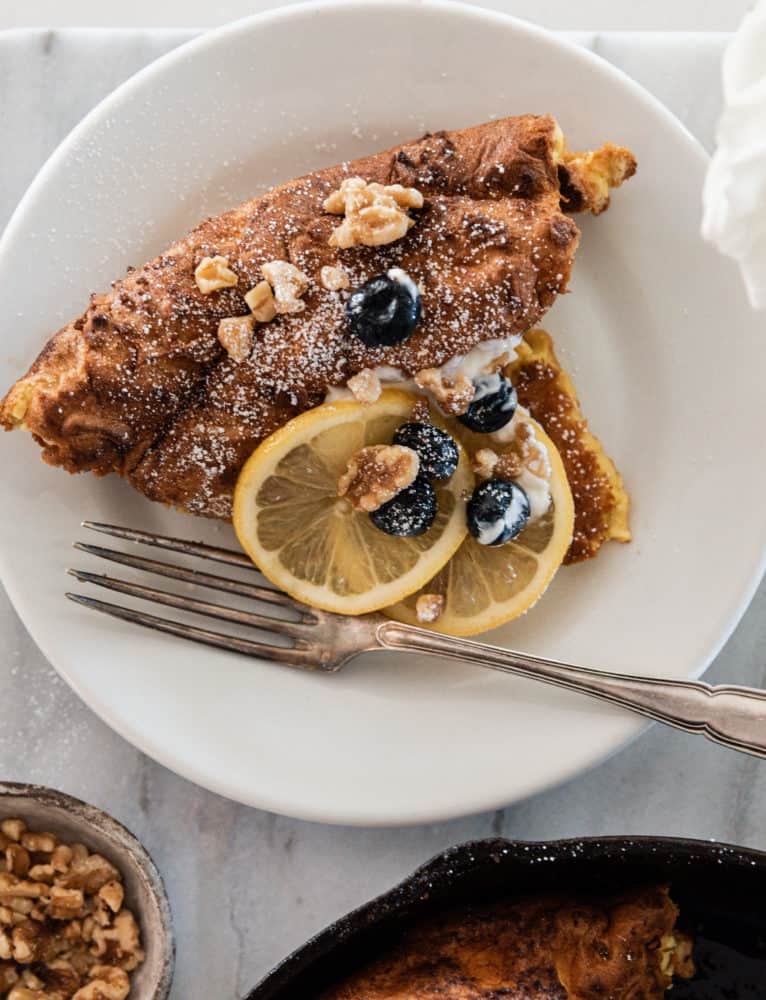 Slice of dutch baby pancake on white plate topped with lemon, blueberries, walnuts, and powdered sugar.
