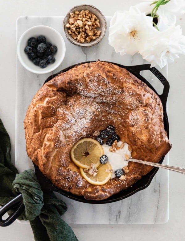 Dutch baby pancake in cast iron skillet topped with yogurt, lemon slices, blueberries, and walnuts - dusted with powdered sugar.