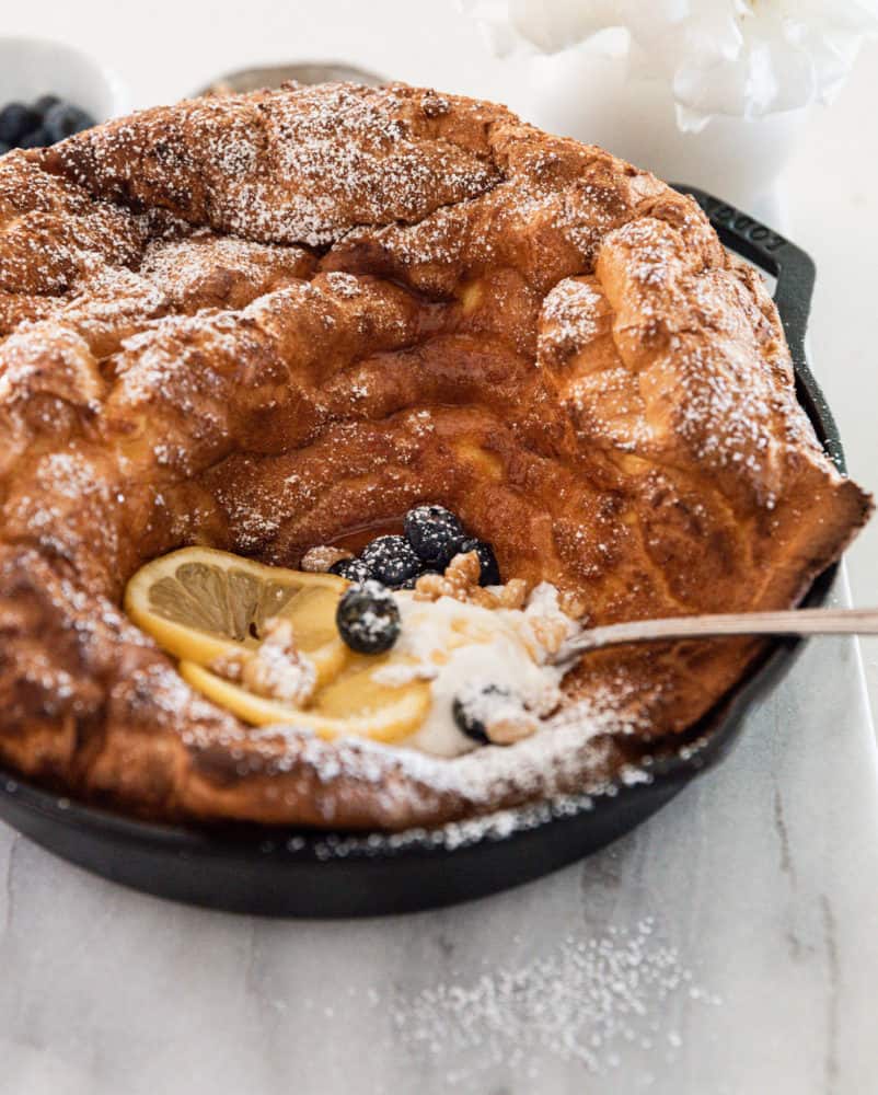 Dutch baby pancake in cast iron skillet topped with yogurt, lemon slices, blueberries, and walnuts - dusted with powdered sugar.