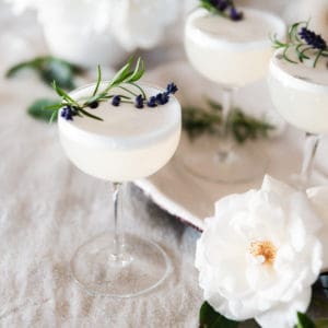 Lavender vodka fizz cocktail on a table with roses and rosemary on a table.