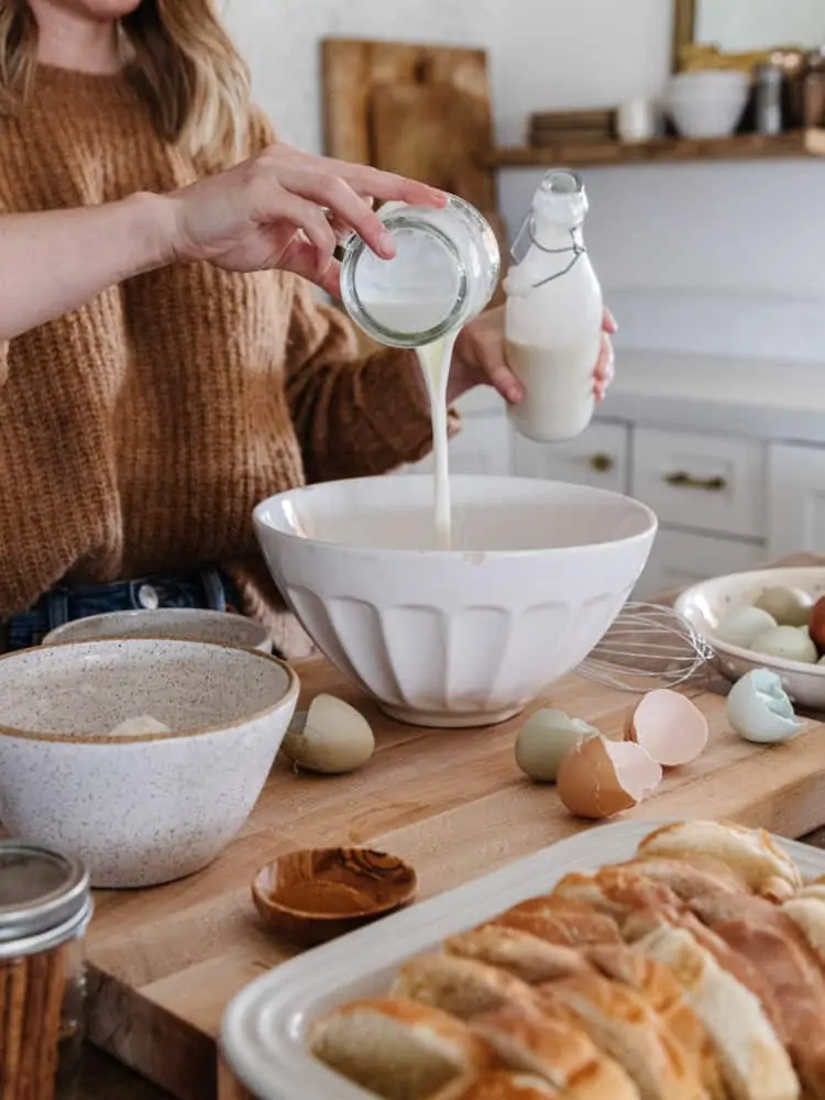 Milk poured into a white bowl on a wood countertop with surrounding ingredients including bread and eggs.
