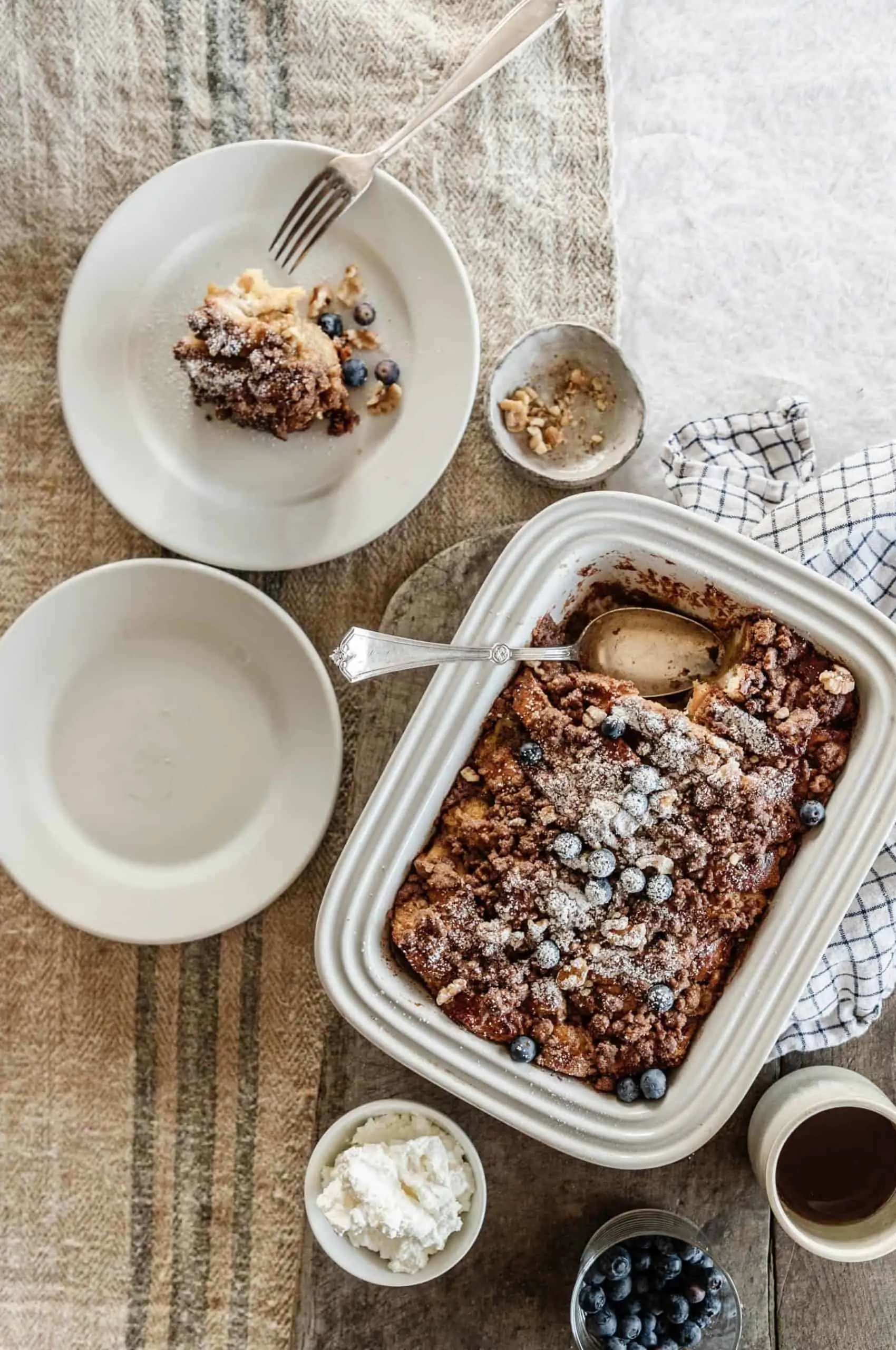 Baked french toast in a white baking dish served with whipped cream, blueberries, maple syrup and topped with powdered sugar.