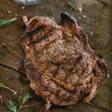 Ribeye steak on wood cutting board with seasoning and rosemary.