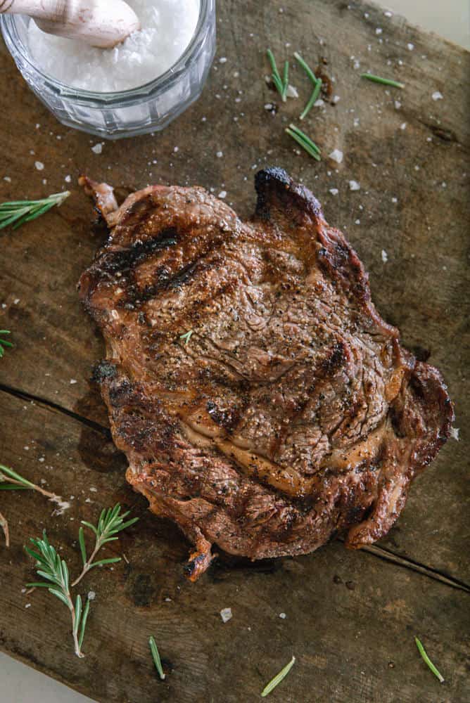 Ribeye steak on wood cutting board with seasoning and rosemary.