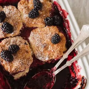 Blackberry cobbler served in a white dish topped with blackberries and powdered sugar.
