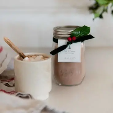 Jar of homemade hot chocolate mix with free printable gift tag and green velvet ribbon on concrete countertop in farmhouse kitchen.