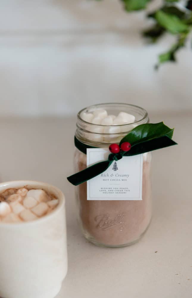 Jar of homemade hot chocolate mix with free printable gift tag and green velvet ribbon on concrete countertop in farmhouse kitchen.