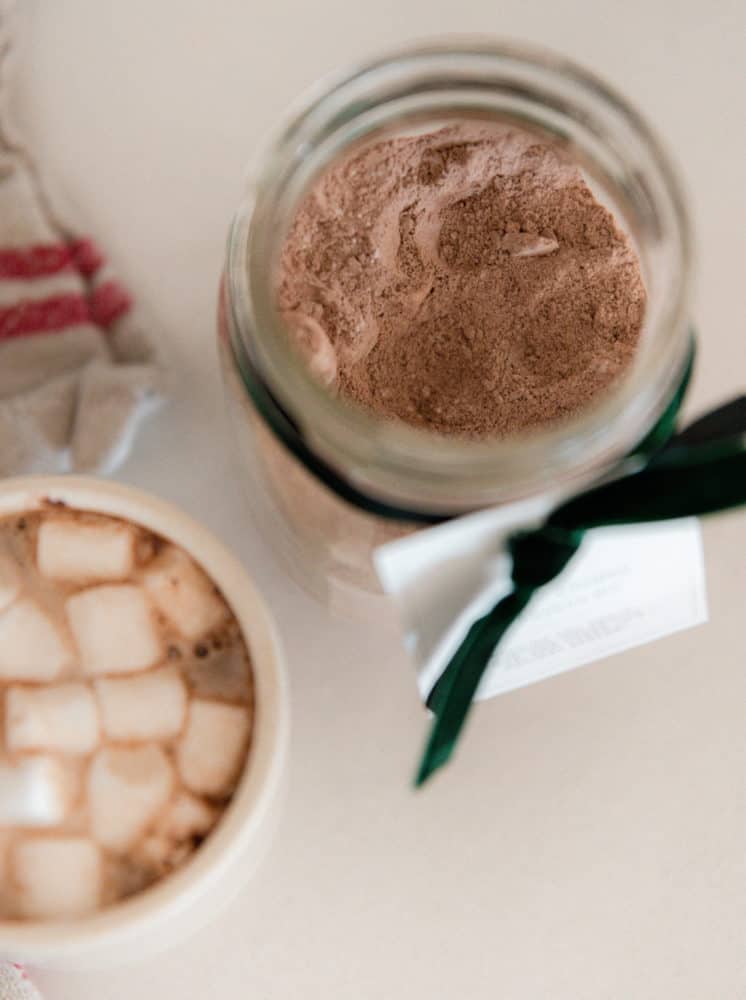 Jar of homemade hot chocolate mix with free printable gift tag and green velvet ribbon on concrete countertop in farmhouse kitchen.