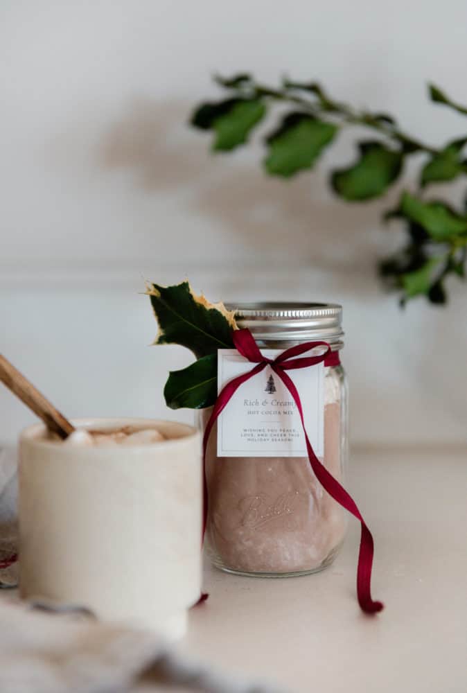 Jar of homemade hot chocolate mix with free printable gift tag and green velvet ribbon on concrete countertop in farmhouse kitchen.