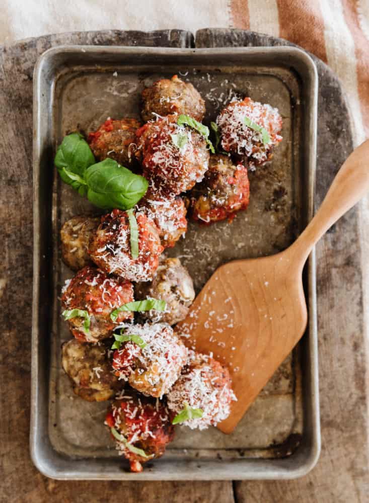 Meatballs topped with cheese and basil on metal pan with a wood spatula ready to serve.