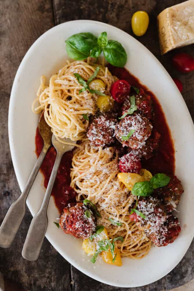 spaghetti and meatballs with basil and cheese on a white oval plate with pasta swirled around a fork