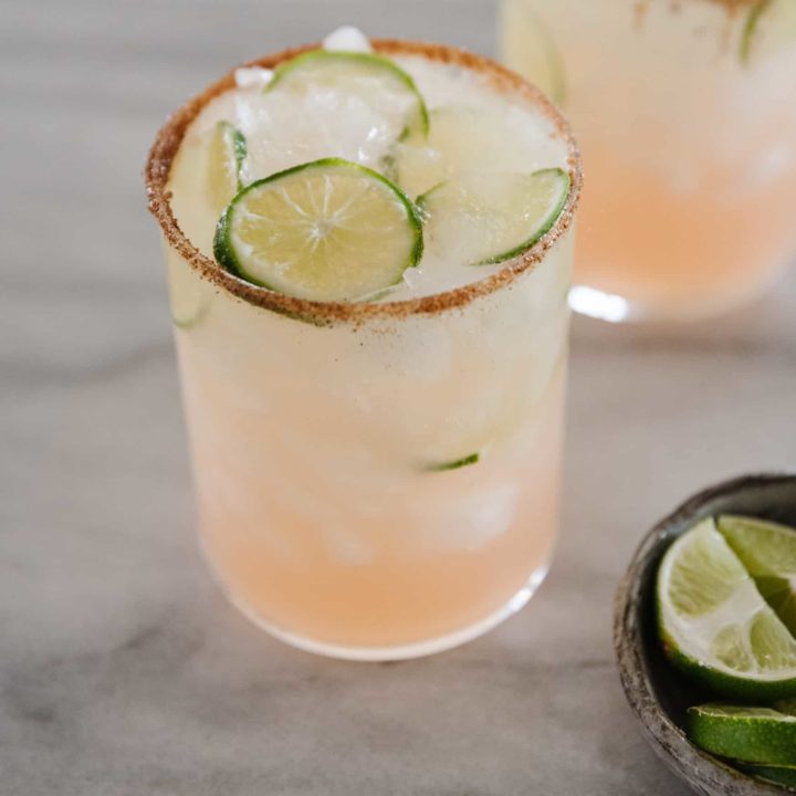 A close up of a Paloma drink with another drink in the background with a bowl of lime wedges