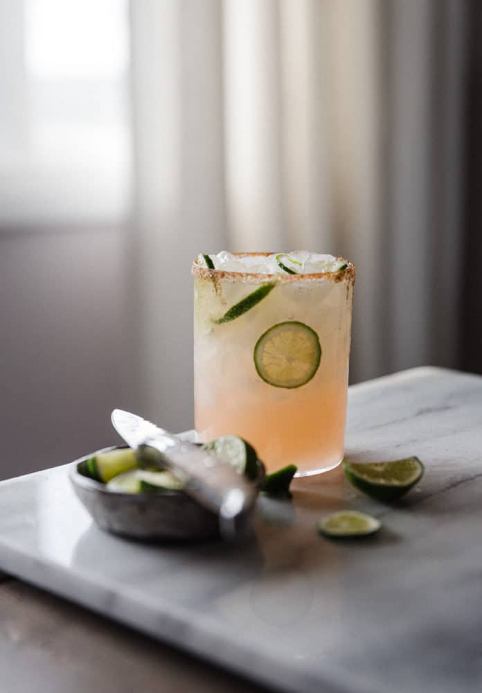 one Paloma drink on a marble cutting board with a knife and cut lime wedges in a bowl