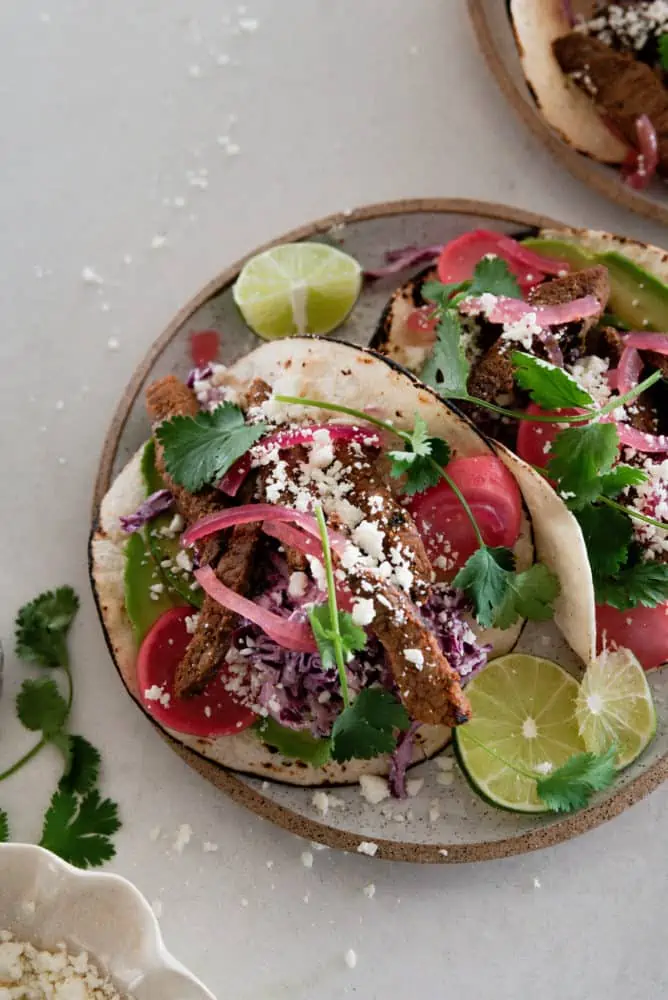 A plate with two steak tacos with cilantro and a lime wedge.