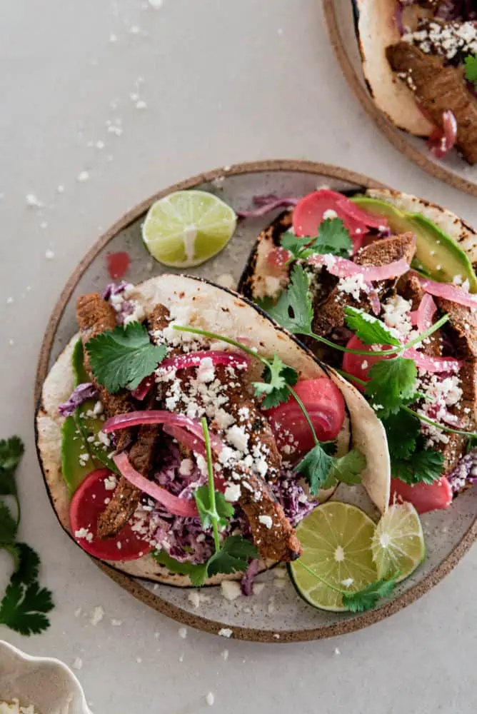 A plate with two steak tacos with cilantro and a lime wedge.