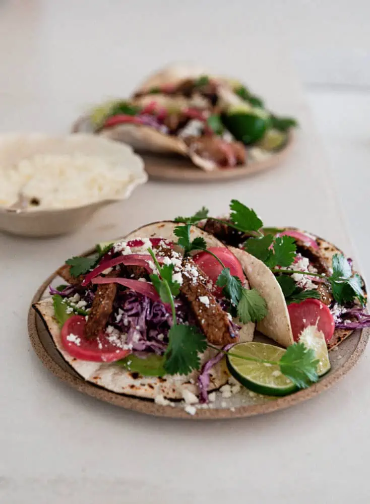 A close-up photo of a plate of steak tacos with a bowl of Cotija cheese and another plate with tacos in the background.