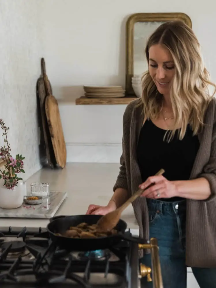 A woman standing at the stove top cooking the steak in a cast iron skillet with a wooden spoon.