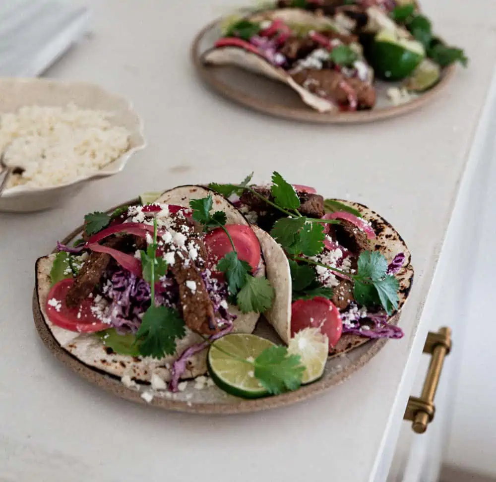 A close-up photo of a plate of steak tacos with a bowl of Cotija cheese and another plate with tacos in the background.