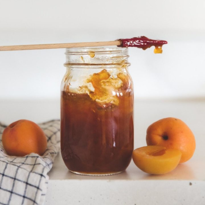 close up of glass jar filled with apricot jam with a spatula resting on top with apricot fruit on a countertop