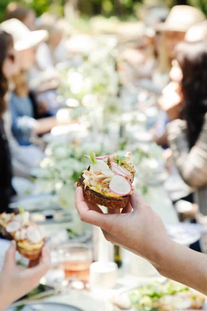 Hand holding a salmon tartine with a blurred background of several people sitting at a long table at an outdoor party.