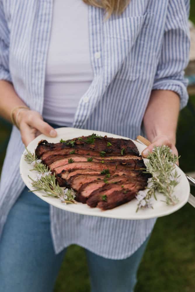 woman holding a white plate with sliced flank steak and rosemary sprigs