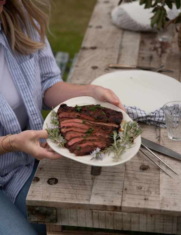 a woman holding a white plate with sliced flank steak at an outdoor rustic table