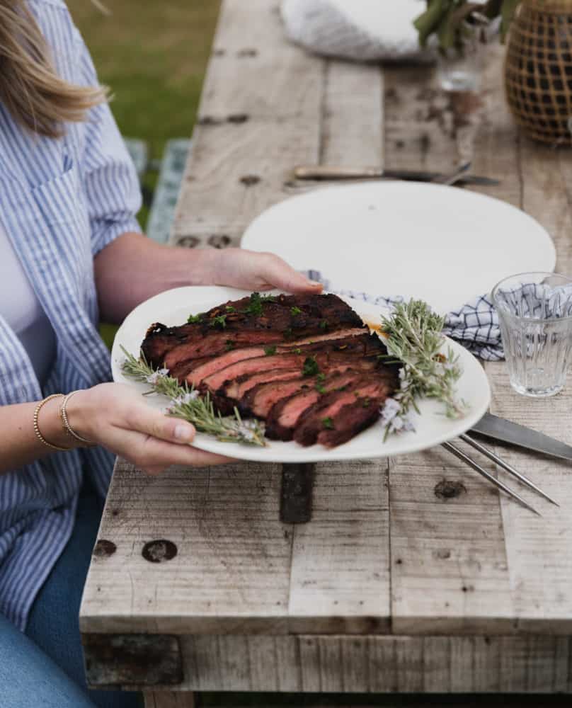 a woman holding a white plate with sliced flank steak at an outdoor rustic table