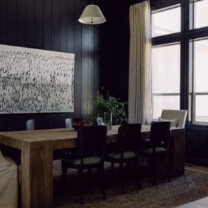angled dining room photo with black walls, rustic chunky dining room table surrounded by chairs with light drapes and artwork