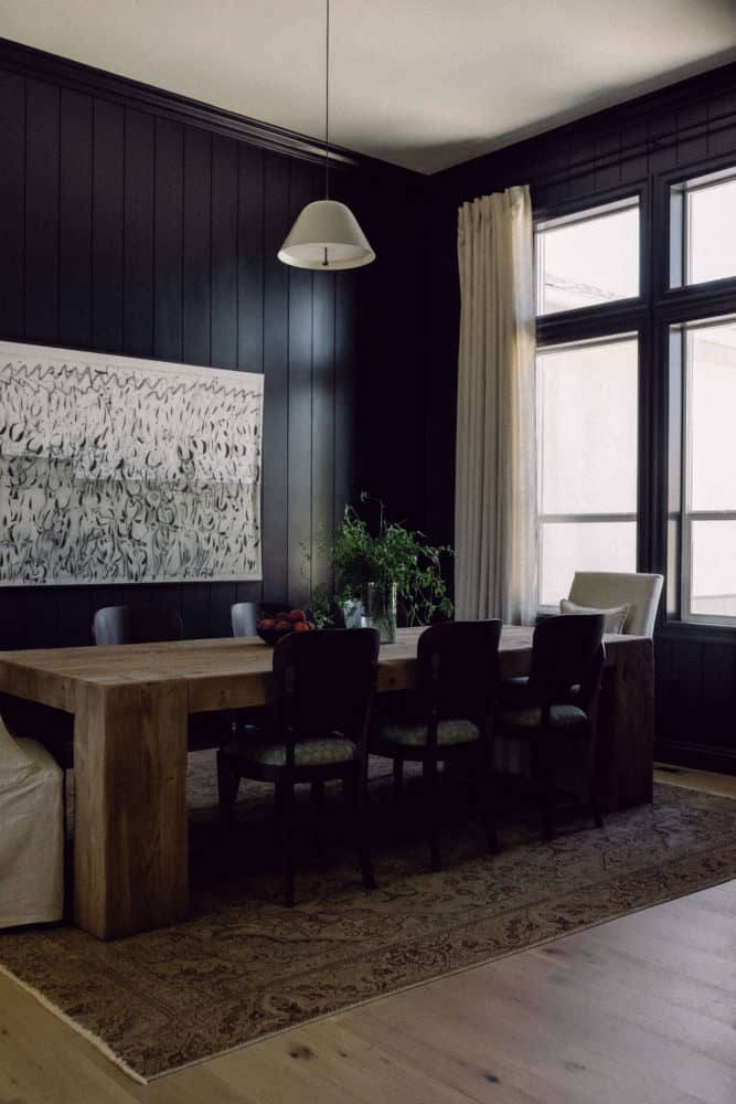angled dining room photo with black walls, rustic chunky dining room table surrounded by chairs with light drapes and artwork