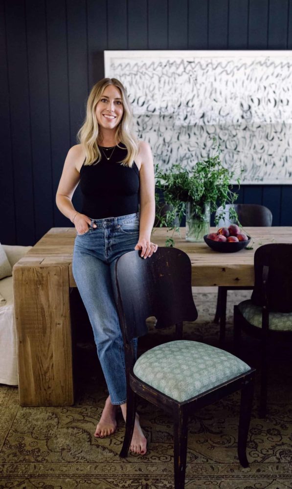 a woman standing beside a wooden dining chair while leaning against a rustic wood table with black walls in the background