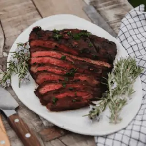 sliced flank steak on white plate on a rustic table with knifes on the table to the left