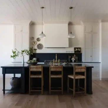 photo of a kitchen with large black island with wicker stools and light grey cabinets with white plaster hood