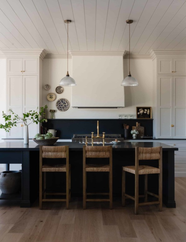 photo of a kitchen with large black island with wicker stools and light grey cabinets with white plaster hood