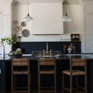 kitchen scene with wicker barstools in front of an all black island, tongue and groove ceiling with plaster hood and light grey cabinets in the background