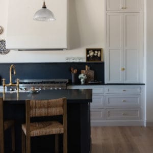 kitchen scene with wicker barstools in front of an all black island, tongue and groove ceiling with plaster hood and light grey cabinets in the background