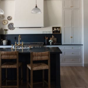 kitchen scene with wicker barstools in front of an all black island, tongue and groove ceiling with plaster hood and light grey cabinets in the background