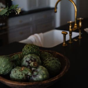 artichokes in wooden bowl on black granite countertop with brass faucet and white sink in background