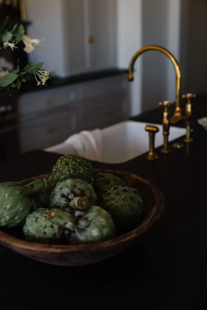 artichokes in wooden bowl on black granite countertop with brass faucet and white sink in background