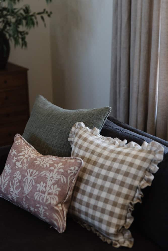 close up photos of three throw pillows on a living room sofa. one is mauve with floral patterns, a beige and white checkered with ruffled edges and a solid sage green pillow