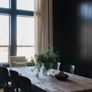 angled view of a dining room with black walls, rustic wood table, and a large window with white linen drapes
